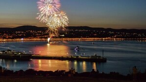 Blick auf das Wasser mit Feuerwerk darüber, einer Anlegestelle und einer Stadtlandschaft im Hintergrund.
