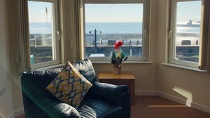 Ein Wohnzimmer mit einem dunkelgrünen Sofa, einem kleinen Holztisch mit Blumen in einer Vase und großen Fenstern, die einen Blick auf die Stadt bieten.