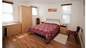 Ein Schlafzimmer mit Holzboden, einem Bett mit gemustertem Überwurf, einem Schreibtisch mit Stuhl und einem Kleiderschrank.