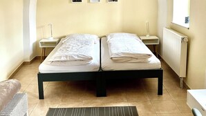 A room with two beds, a small table, a chair, a radiator, and three framed pictures on the wall.