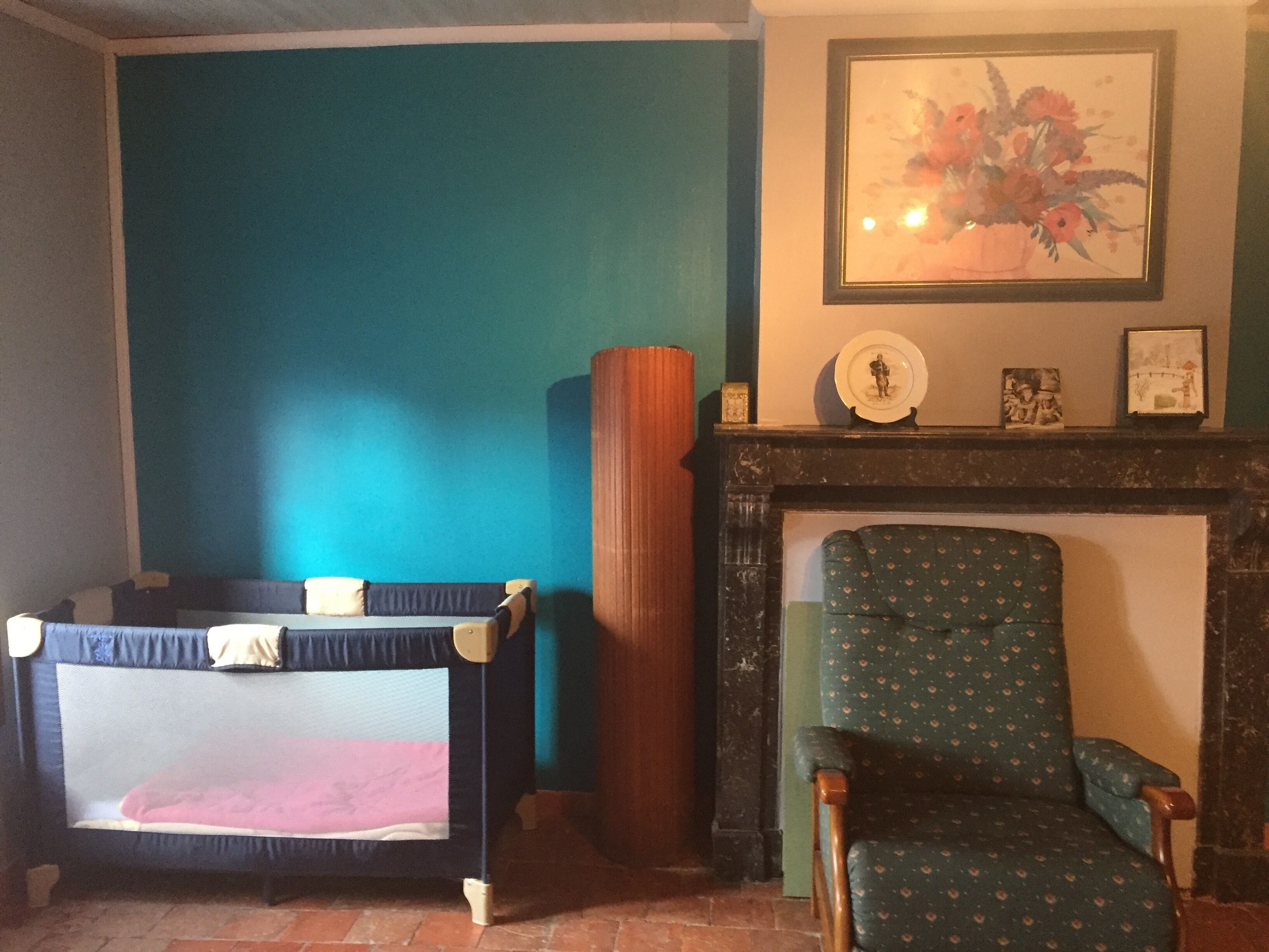 Een kamer met een ledikant, een groene fauteuil, een open haard en een lijst met een bloemistisch schilderij aan de muur.