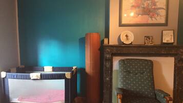 Een kamer met een ledikant, een groene fauteuil, een open haard en een lijst met een bloemistisch schilderij aan de muur.