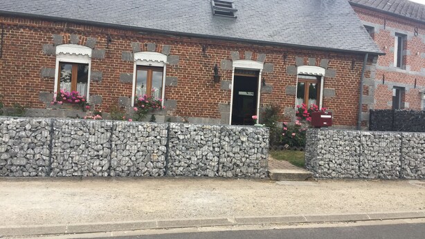 Una casa de ladrillo con un muro de piedra en primer plano, una puerta negra y ventanas con flores rosadas.