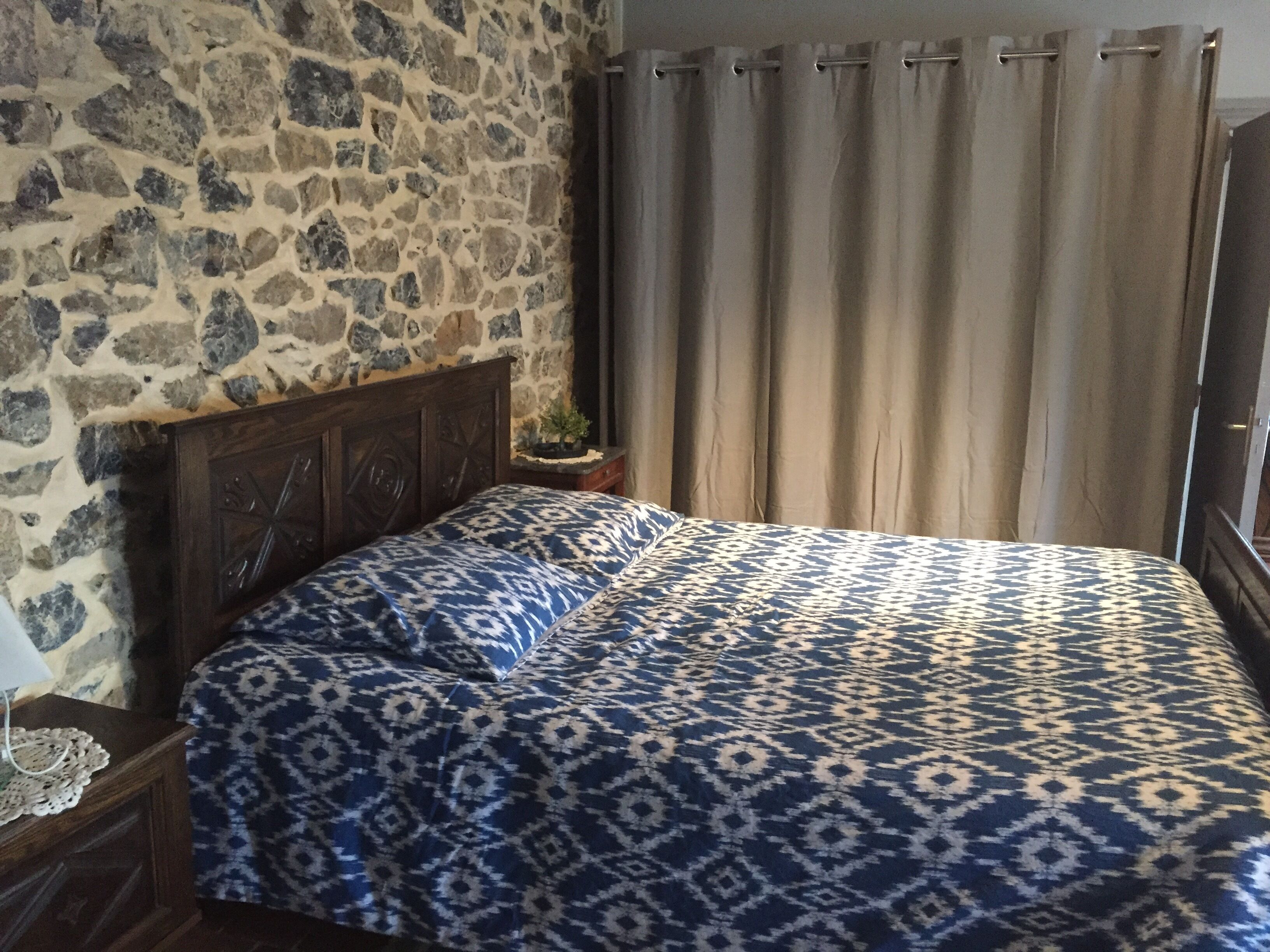 A bedroom with a stone wall, a wooden headboard, a patterned bedspread, and a curtain.