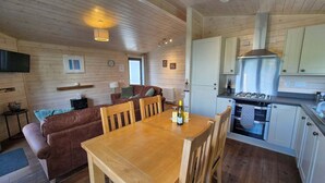 A cozy cabin interior with a wooden dining table, a sofa, and a kitchen area with a stove and cabinets. - Lilybank Lodges - 5 Star Self-Catering (Isle of Man)