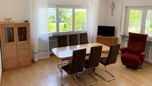 A room with a wooden dining table, four chairs, a red armchair, a television, and a cabinet with glass doors. - Bright apartment (ground floor) with garden and terrace. Experience Hegau, Lake Constance, Rhine Falls! (Tengen)