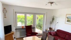 A modern living room with a wooden dining table, red and brown upholstered chairs, a red sofa, a television on a wooden cabinet, and a large window with a view of greenery. - Bright apartment (ground floor) with garden and terrace. Experience Hegau, Lake Constance, Rhine Falls! (Tengen)