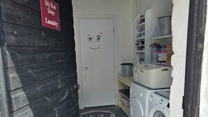 Un espace dédié à la lessive, avec une porte sur laquelle est inscrit « Lessive Lily Bomb », un lave-linge, un étendoir et des étagères remplies de livres et de produits.