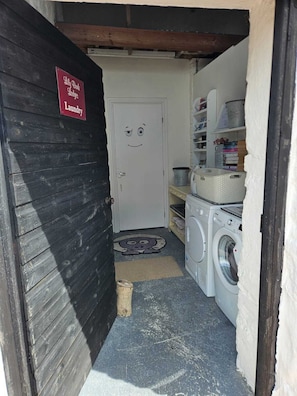 A laundry area with a door marked 'Lily Bomb Laundry,' a washing machine, a drying rack, and shelves with books and supplies. - Lilybank Lodges - 5 Star Self-Catering (Foxdale. Isle of Man)