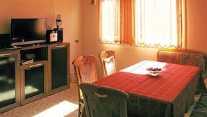 Una habitación con televisión, una mesa pequeña con mantel rojo, dos sillas y una ventana con cortinas.