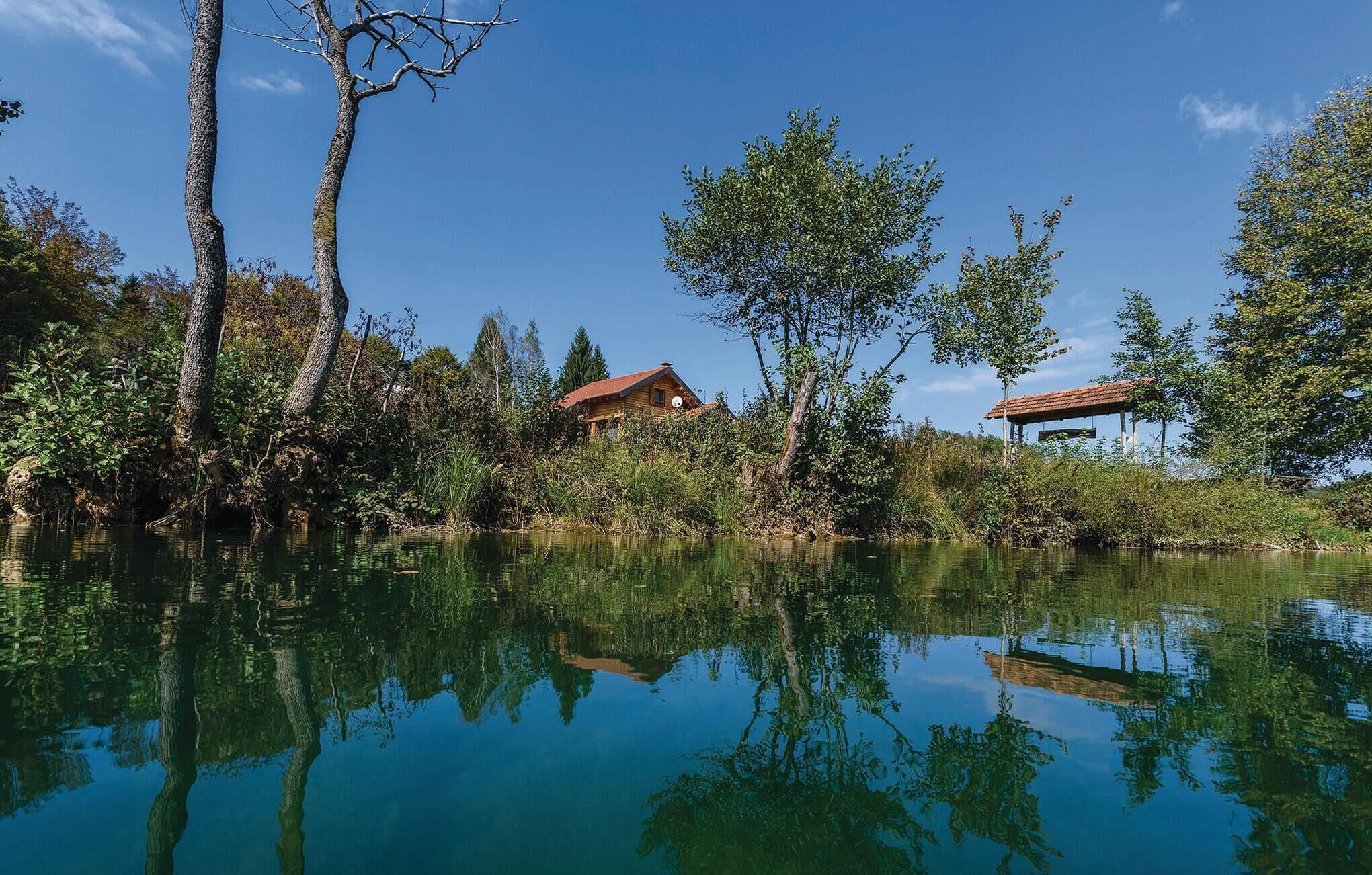 Um cenário tranquilo à beira de um lago, com uma cabana de madeira e um gazebo, cercado por vegetação exuberante e árvores refletidas na água calma.