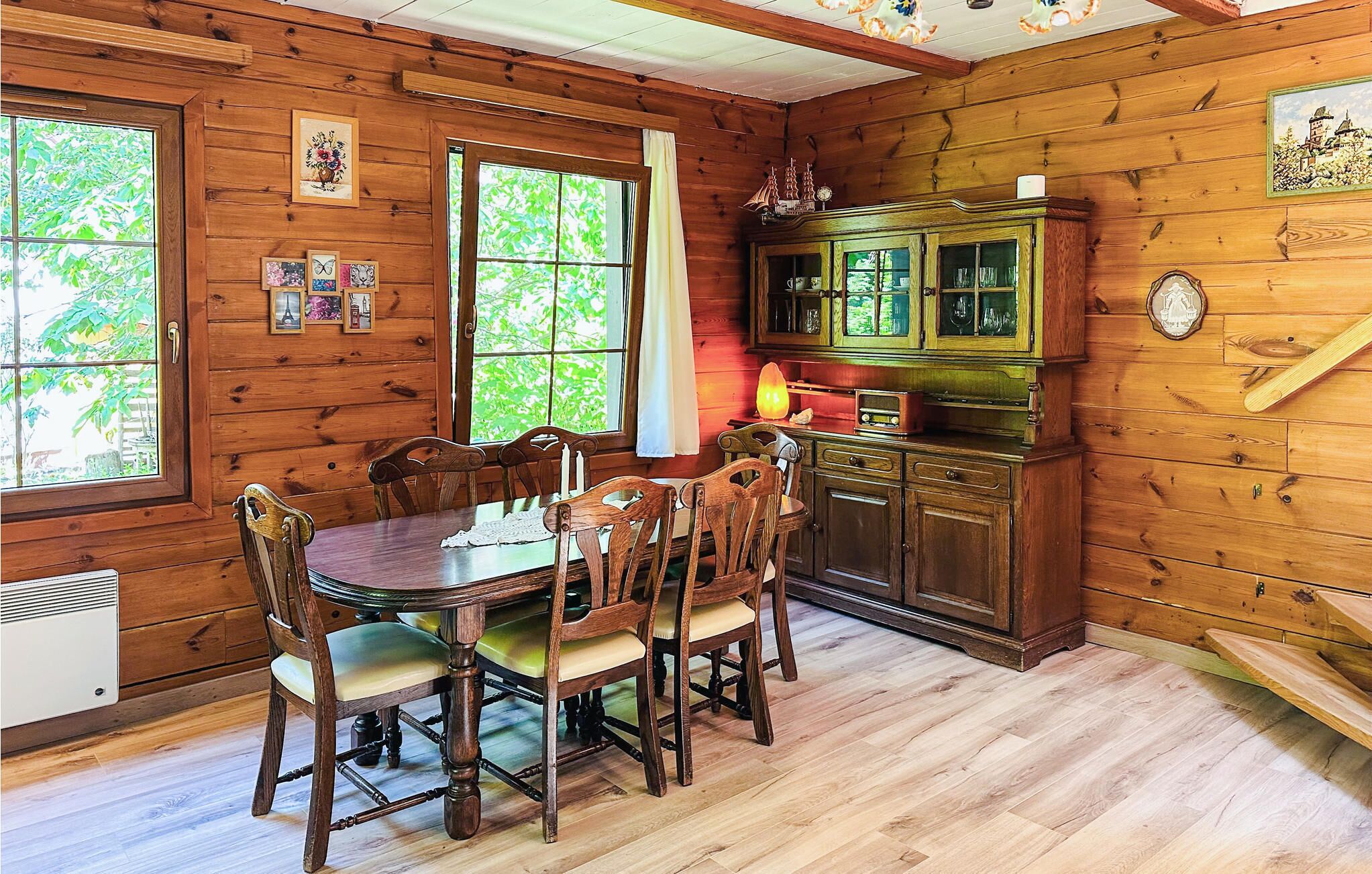 Interior de una cabaña de madera con una mesa de comedor y sillas, un armario con puertas de cristal y una ventana que da a vistas de vegetación.