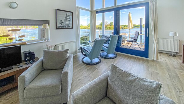 Un salon moderne avec une grande fenêtre donnant sur un lac, des canapés gris, un téléviseur et un tableau encadré accroché au mur.