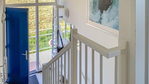 Un escalier avec une rampe blanche mène à un balcon. Une porte bleue est visible sur la gauche, et un tableau représentant un phare est accroché au mur sur la droite.