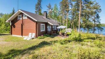 Une cabane en bois dotée d’une terrasse, entourée d’arbres et bordée par un lac.