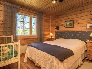 A wooden cabin bedroom with a bed, a nightstand, a lamp, a crib with a stuffed animal, and a window with curtains.