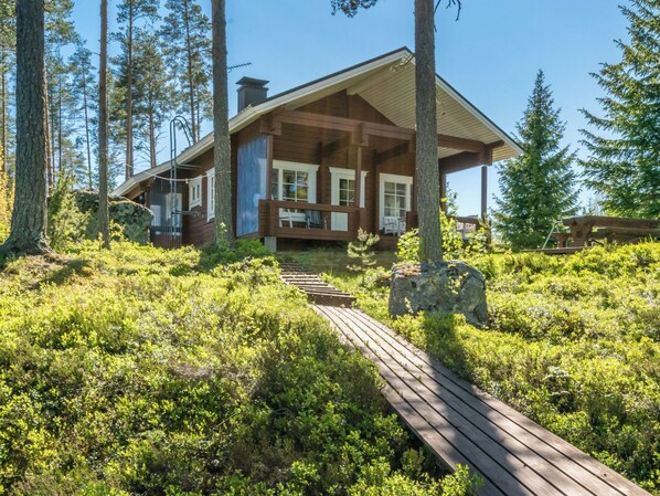 A cabin with a wooden deck leading to a covered porch, surrounded by dense forest.