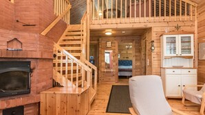 A wooden cabin interior with a fireplace, a staircase, a bed, and a kitchen area.