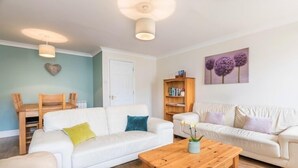 A living room with two white sofas, a wooden coffee table, a bookshelf, and a heart-shaped decoration on the wall. - Beautiful 4* Self-Catering, by Stunning Cliffs & Bays - Three Bedroom Cottage (Guernsey)