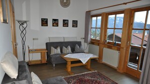 Un salon avec un canapé gris, une table basse en bois, un tapis à motifs et une grande fenêtre donnant sur un paysage montagneux.