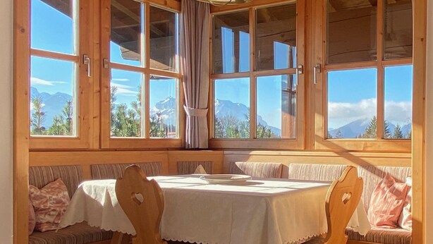 Une salle à manger en bois avec une table et des chaises, de grandes fenêtres avec des rideaux et une vue sur les montagnes.
