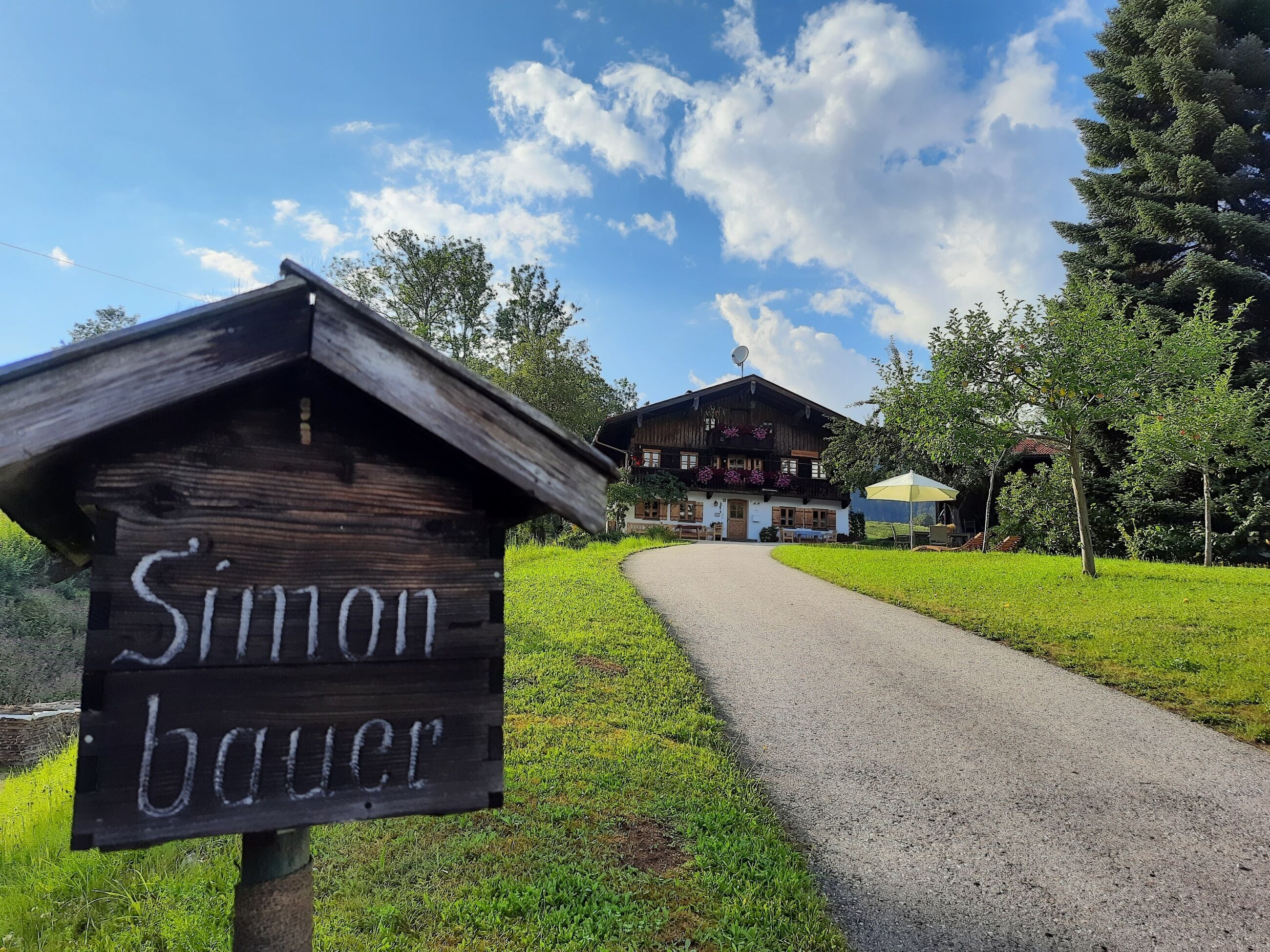 Ein hölzernes Schild mit der Aufschrift "Simon Bauer", ein Weg, der zu einem Gebäude mit geneigtem Dach führt, umgeben von Grünpflanzen und Bäumen.