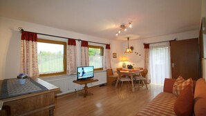 A cozy living room with a wooden TV stand, a sofa, a dining table with chairs, and a window with curtains. - Predigtstuhl vacation apartment in a rural setting, 55 m², 1 bedroom (Bayerisch Gmain)