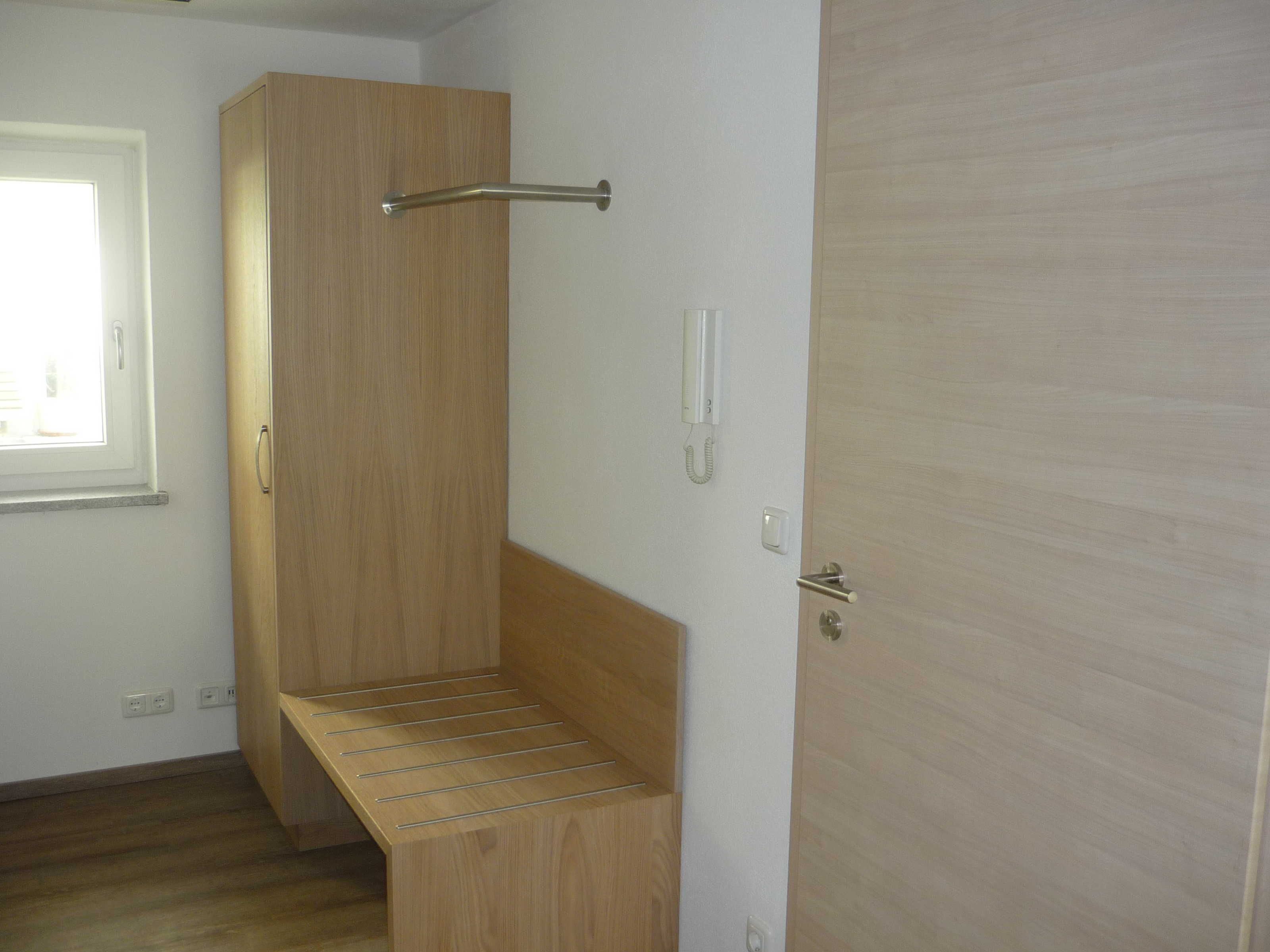 A room with a wooden wardrobe, a bench, a door with a handle, and a light switch.