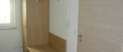 A room with a wooden wardrobe, a bench, a door with a handle, and a light switch.