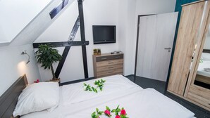 Ein Schlafzimmer mit schrägen Wänden, einem an der Wand montierten Fernseher, einer Holzkommode und einem Bett mit weißen Leinen und rosa Blumen.