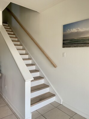 Eine Treppe mit Holzstufen und weißen Geländern, die zu einer Zwischenebene führt, auf der ein an der Wand befestigtes Bild einer Strandlandschaft zu sehen ist.