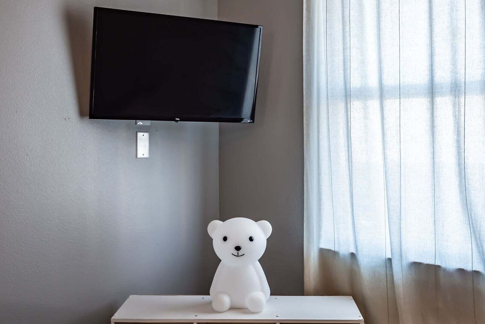 Una habitación con un televisor montado en la pared, un objeto con forma de oso blanco sobre una mesa y una ventana con cortinas.