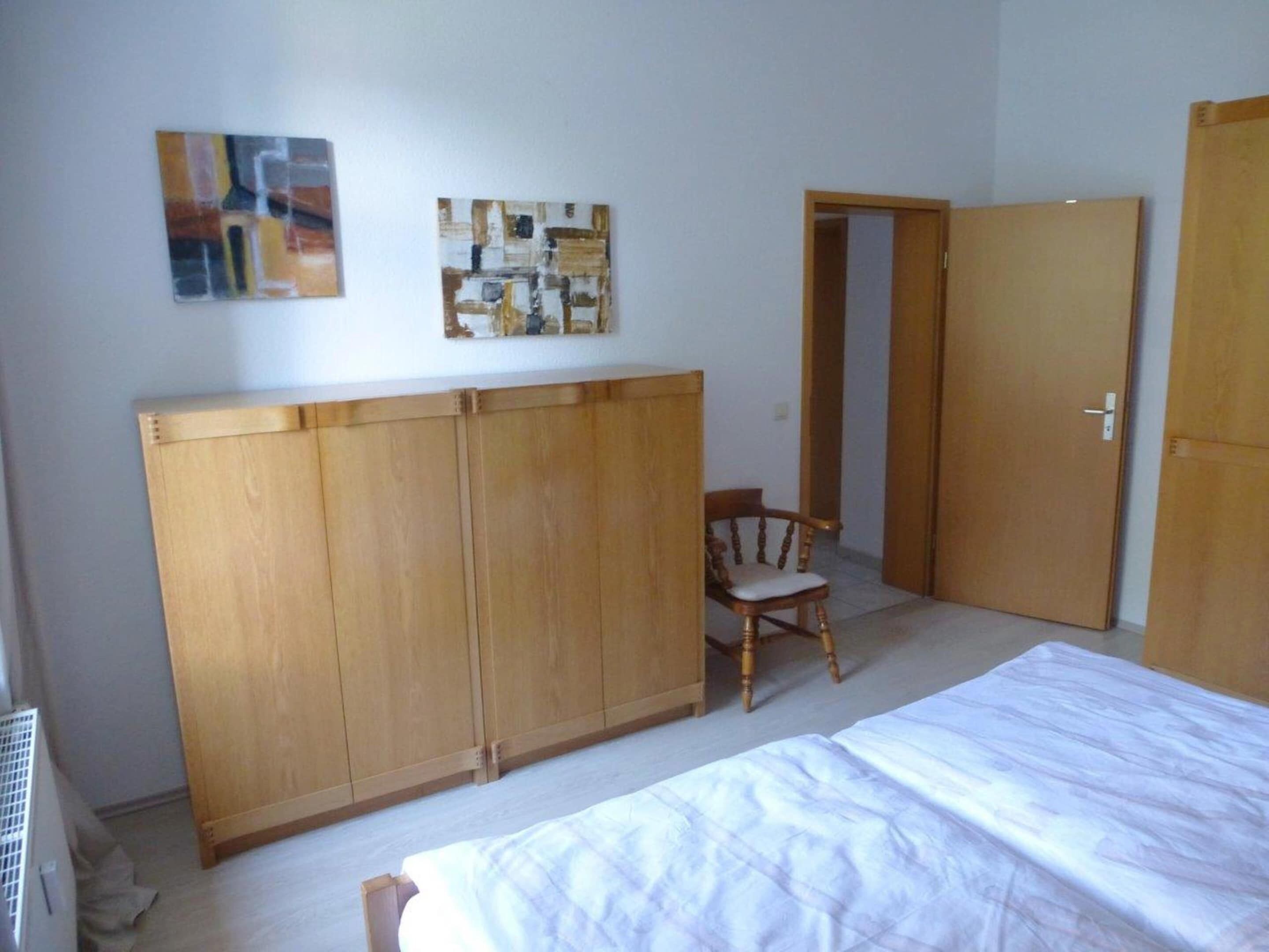 Ein Schlafzimmer mit einem Bett, einem Holzschrank, zwei Wandbildern, einem Sessel und einer Tür.