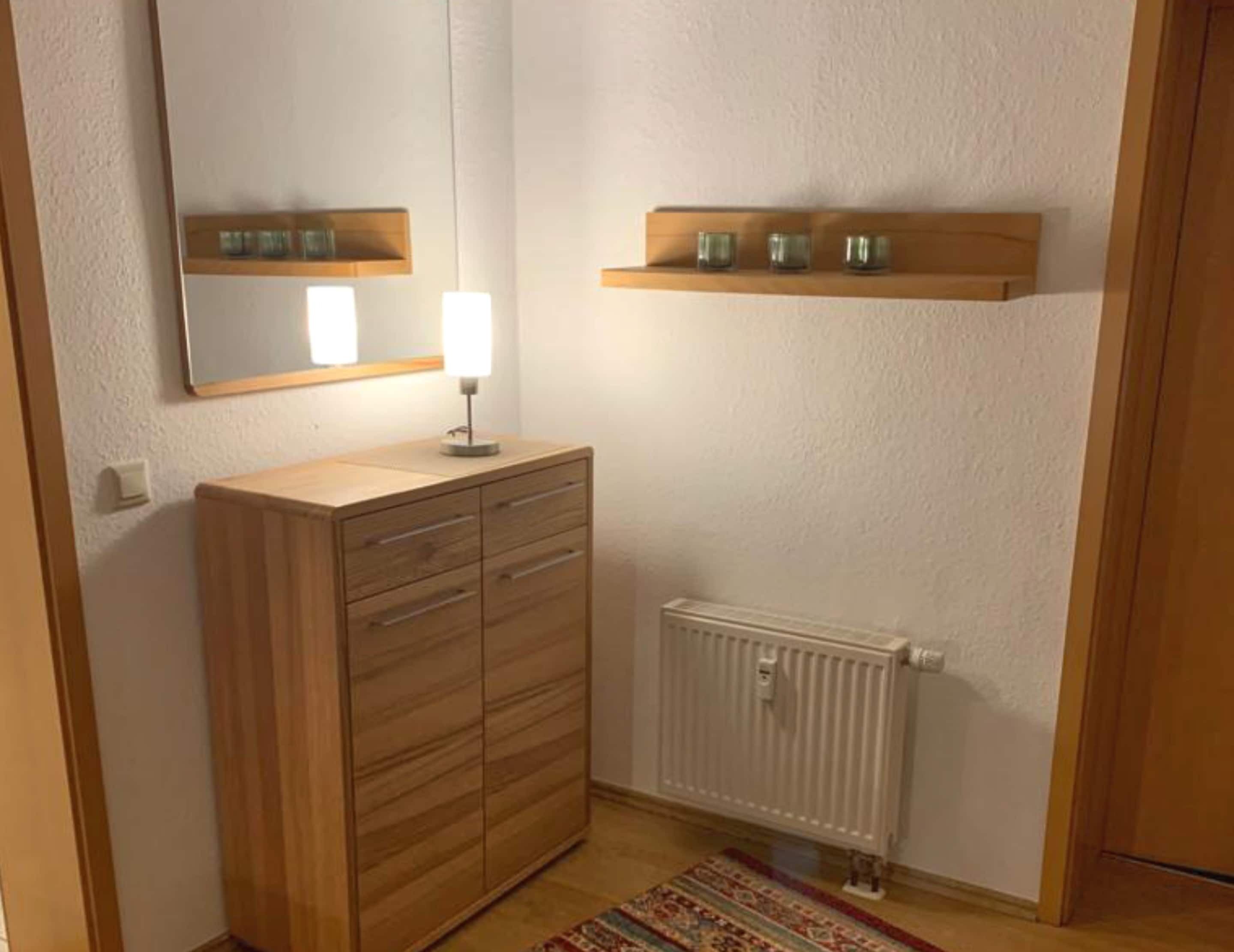 A small, well-lit bathroom with a wooden cabinet, a mirror, a shelf with glass jars, and a radiator.
