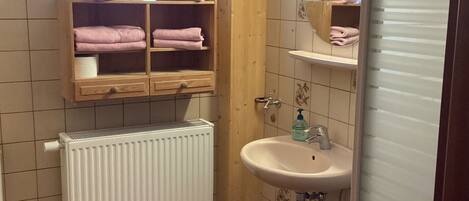 A small bathroom with a white radiator, a sink, and a shelf with folded towels.