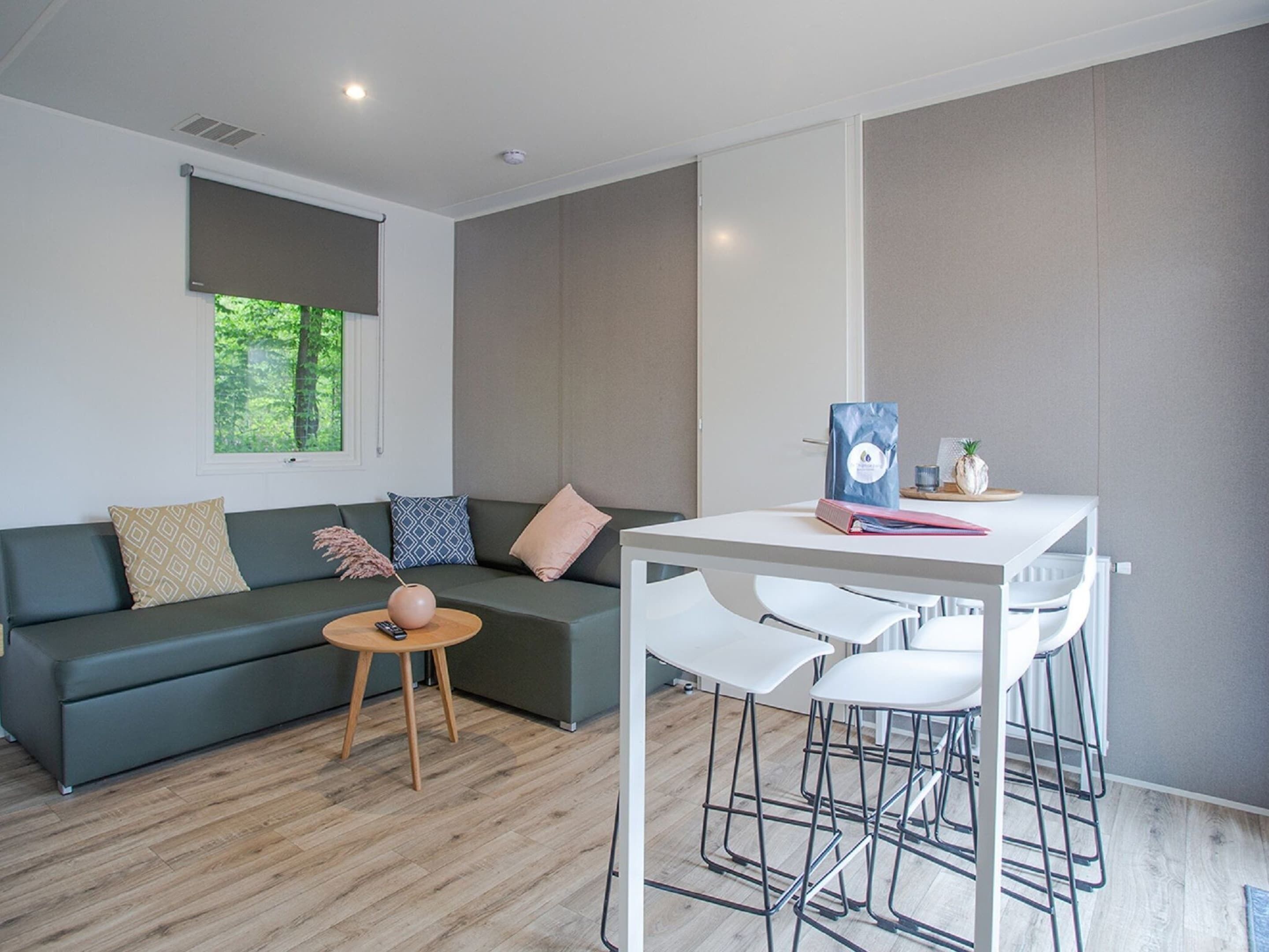 A modern living room with a grey sofa, a small round table, and a white dining area with bar stools.