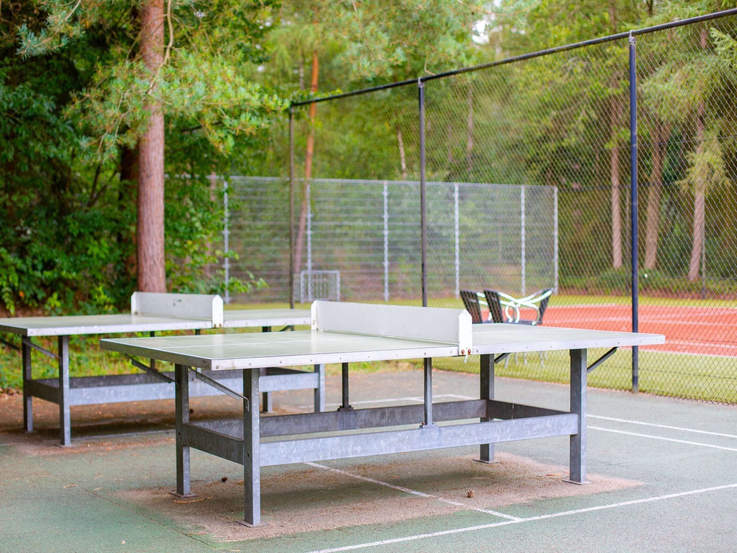 Een tennisbaan met twee witte tafels en bankjes, omringd door een gaashek en bomen.