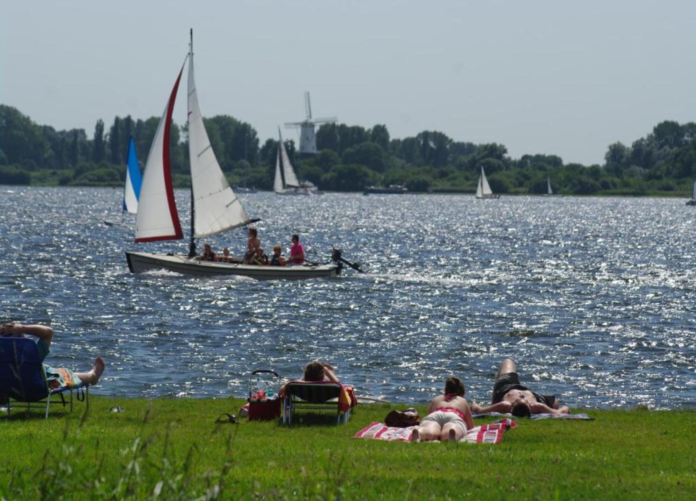 A lake with sailboats and people relaxing on the shore.
