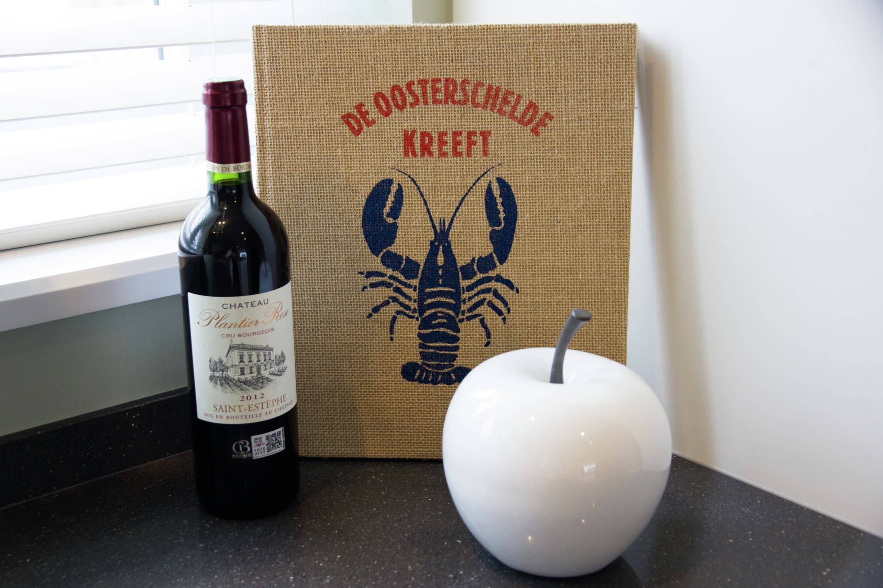 A bottle of Chateau Plantier Ros Cru Bordeaux 2012 wine, a lobster graphic, and a white apple on a countertop.