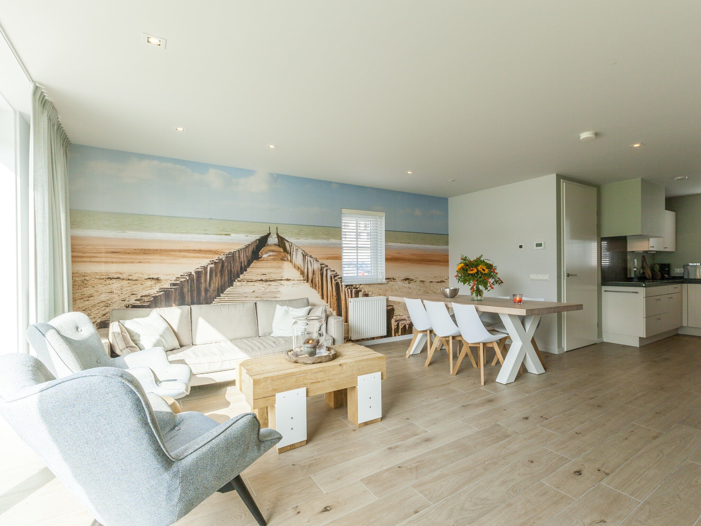 Una sala de estar moderna con un mural de playa, un comedor con sillas blancas y una cocina con armarios blancos.