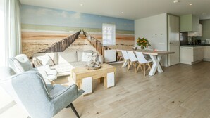 A modern living room with a beach mural, a dining area with white chairs, and a kitchen with white cabinets. - Villa in Cadzand Near Beach & Dunes (Cadzand-Bad)