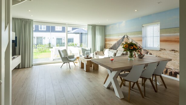 A spacious dining area with a wooden table and chairs, a view of the beach through large windows, and a beach-themed mural on the wall. - Villa in Cadzand Near Beach & Dunes (Cadzand-Bad)