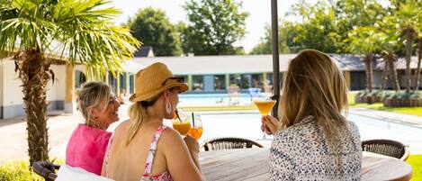 A group of people sitting at an outdoor table under a yellow umbrella, with a pool and greenery in the background.