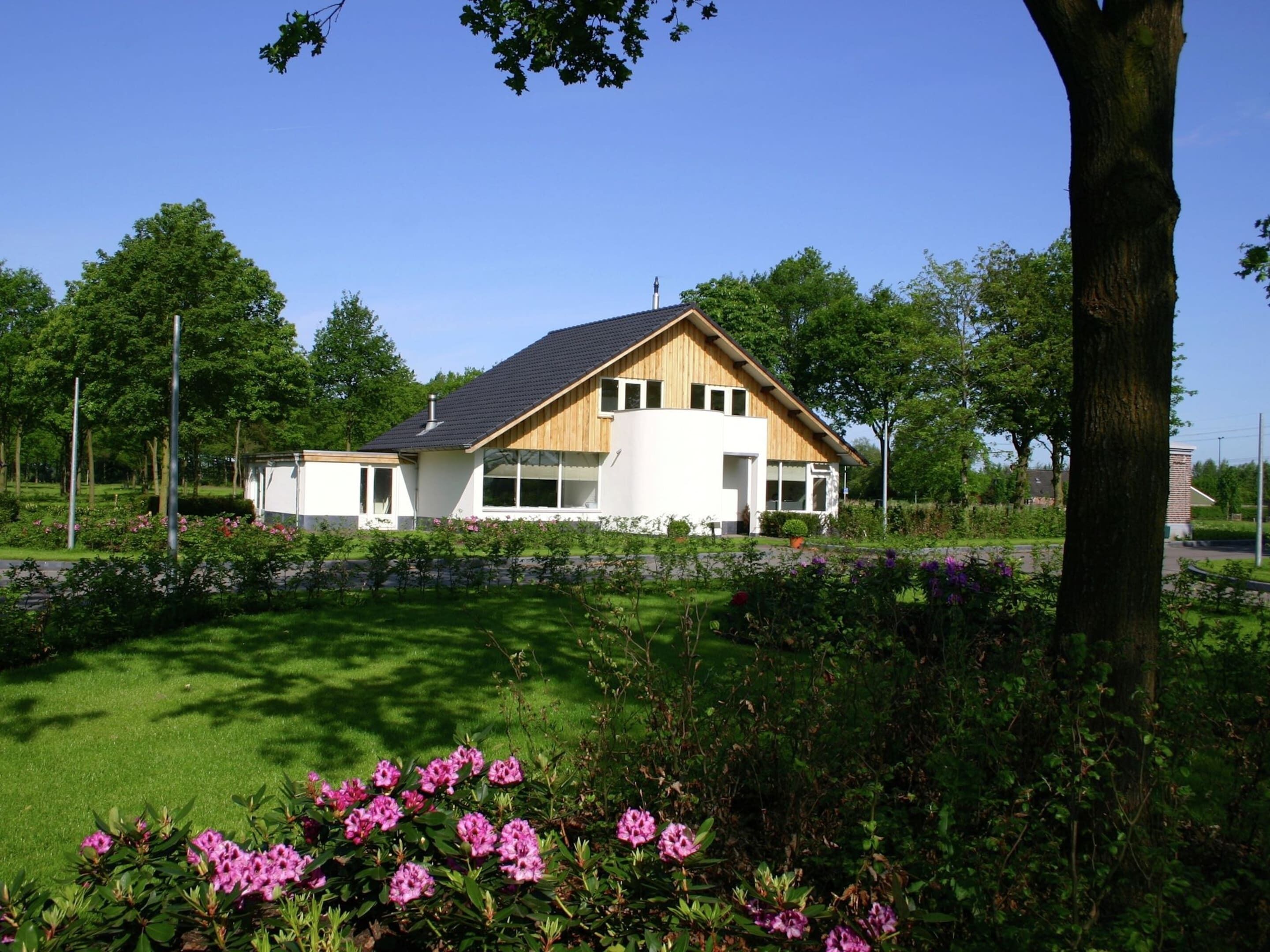 A modern two-story house with a sloped roof, surrounded by a well-maintained garden with pink flowers.
