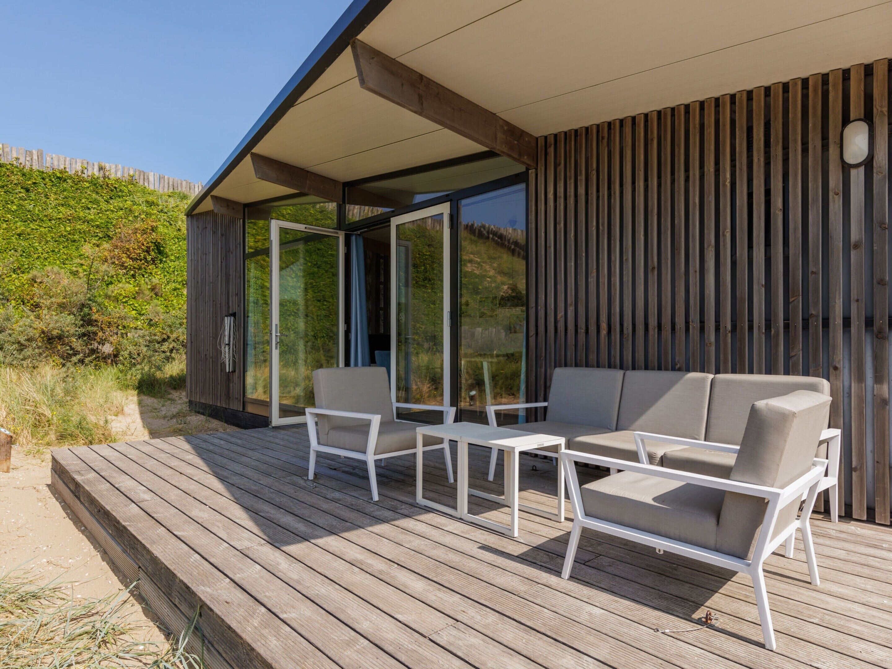 A modern outdoor seating area with a wooden deck, a glass door leading to a covered patio, and a wooden fence in the background.