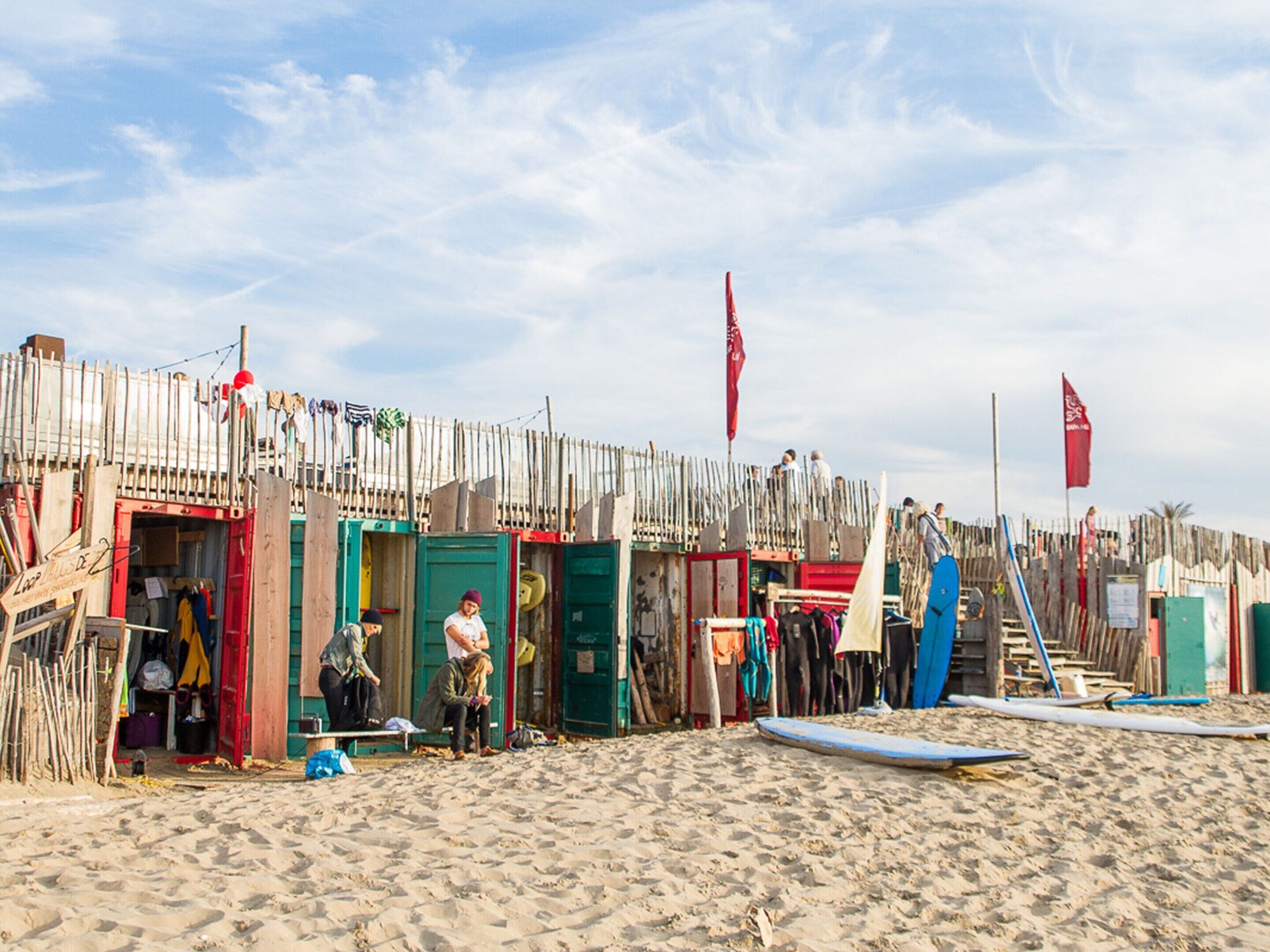 A beach with wooden structures, some with doors and windows, and a few people sitting or standing.