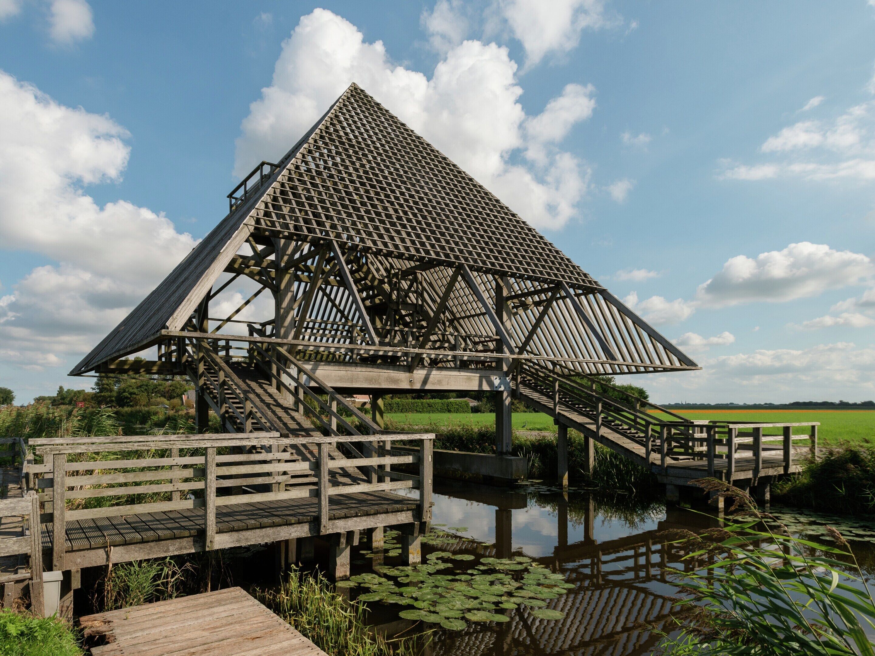 A wooden observation tower with a triangular roof and a staircase leading up to it, situated over a body of water with lily pads.