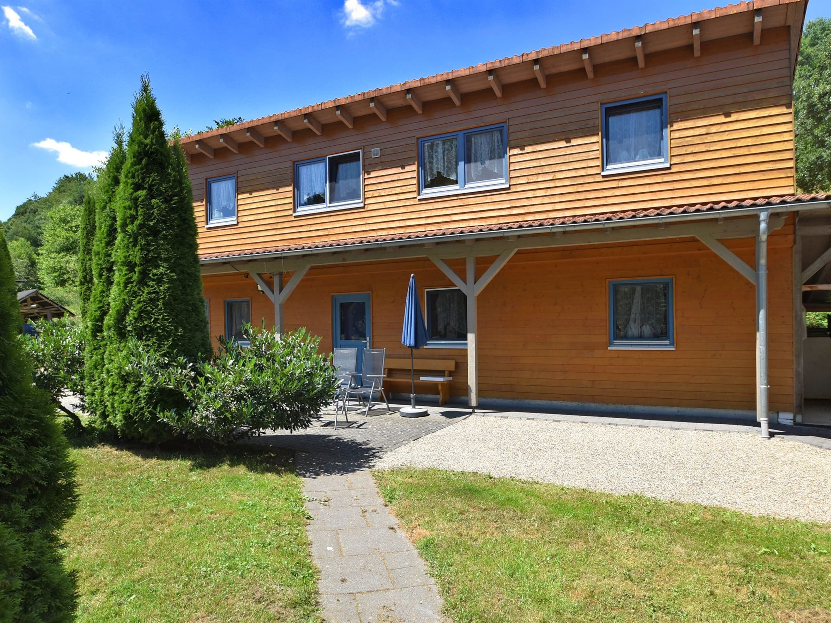 A two-story wooden house with a covered patio, a gravel driveway, and a small garden area.