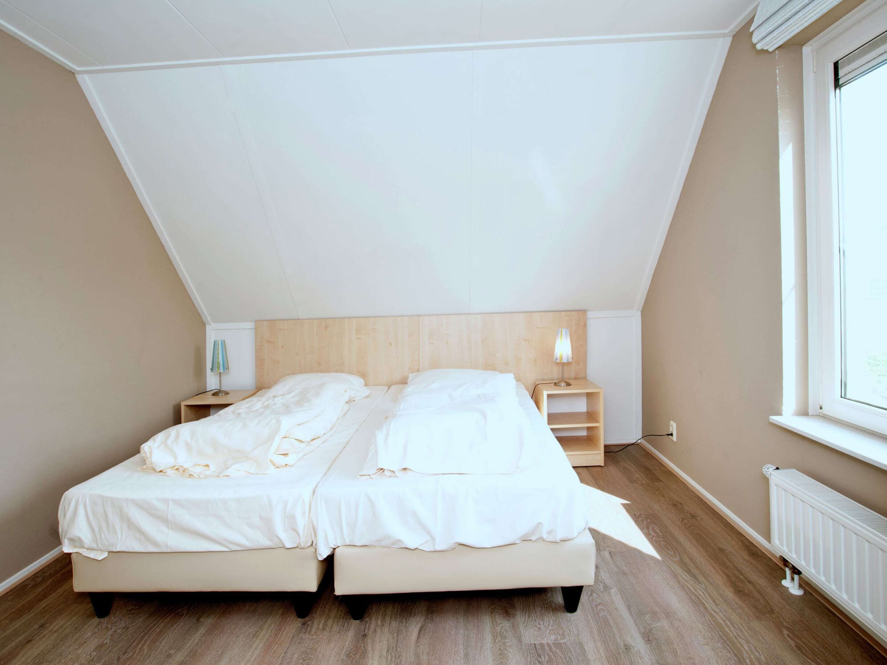 A bedroom with two beds, a wooden headboard, bedside tables, a lamp, a radiator, and a window.