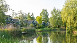 Uma fileira de casas tradicionais à beira de um lago tranquilo, com ervas altas e árvores ao fundo.